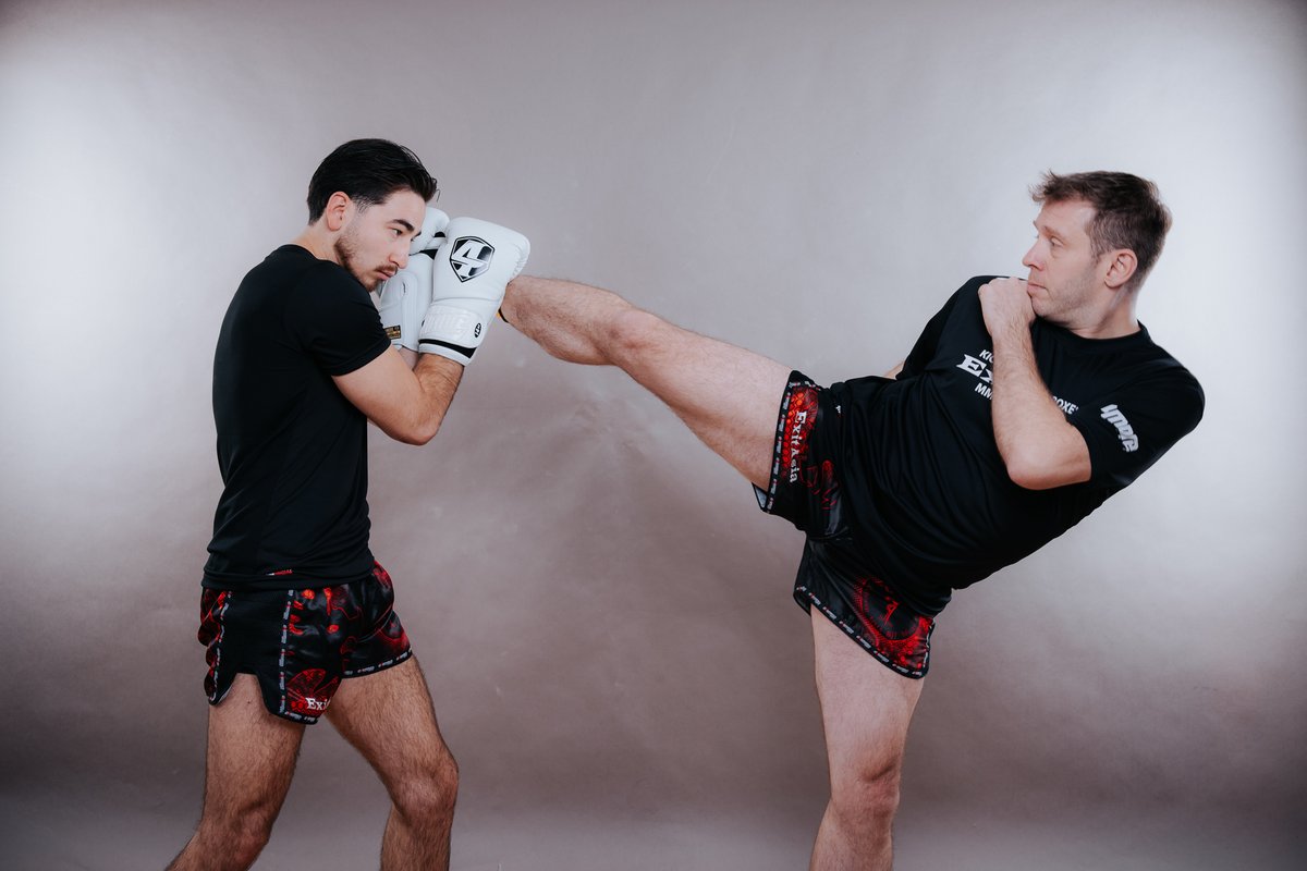 Zwei Kickboxer in schwarzen Shirts: Einer schlägt einen hohen Halbkreistritt gegen die Doppeldeckung des Partners, die eigene Faust bleibt am Kinn